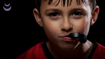 Kid with brown eyes looking directly into the camera with a mouthguard coming out of his mouth.