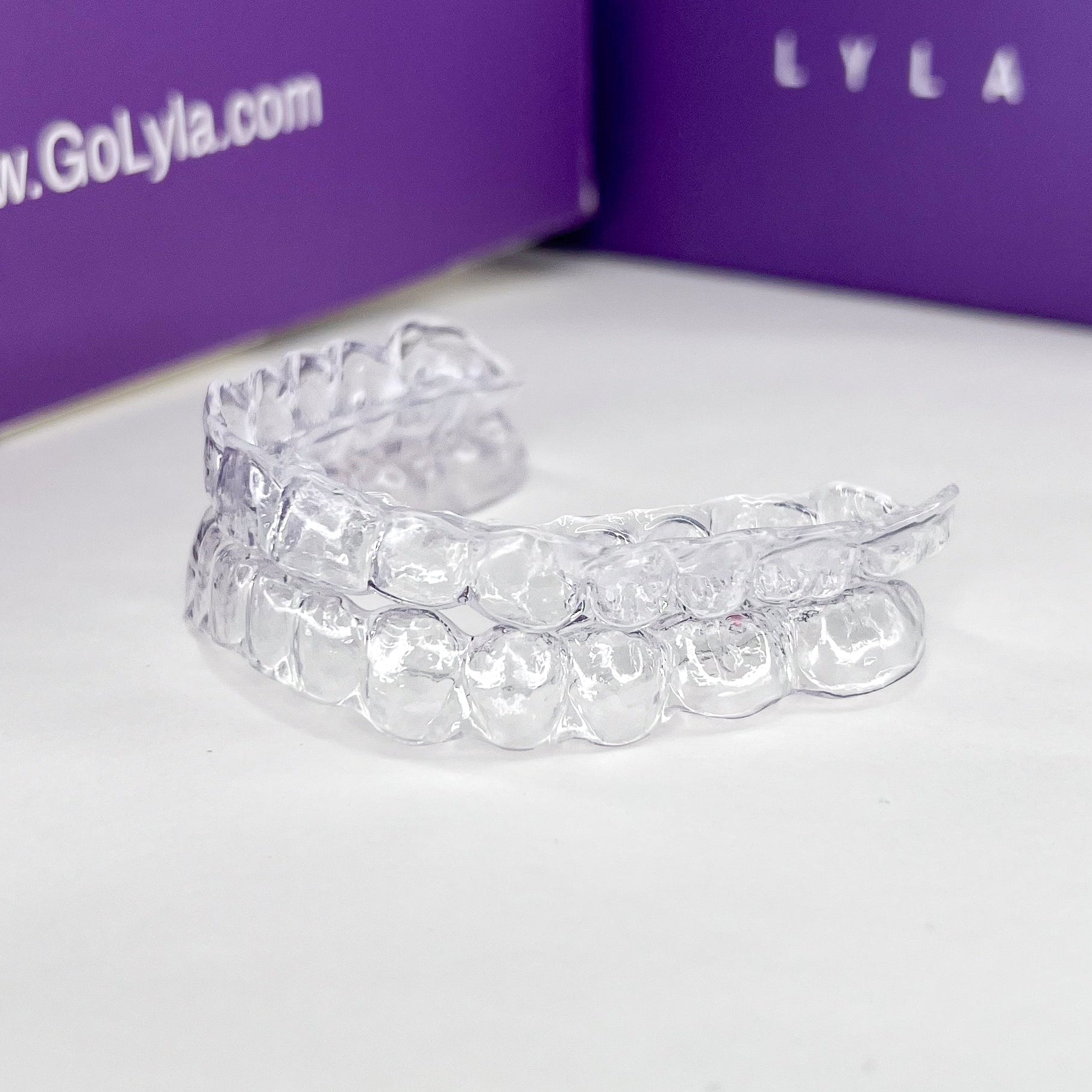Clear dental aligners on a white surface with a purple Lyla box in the background.